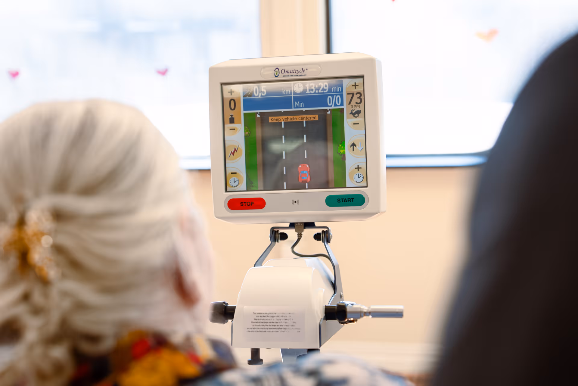 An elderly person with gray hair is using a stationary exercise bike with a digital screen displaying a virtual driving game. The screen shows a road with a red car and instructions to keep the vehicle centered.