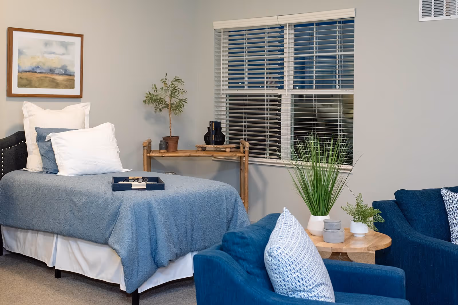 A cozy bedroom with a single bed covered in a blue quilt and white pillows. A small wooden table with decorative items and a potted plant is placed near a window with white blinds. In the foreground, there is a blue armchair with a patterned cushion and a round wooden side table holding two small plants and decorative containers.