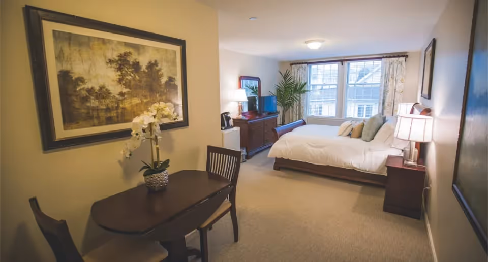 A cozy bedroom in a senior living facility with a large bed adorned with pillows, two bedside tables with lamps, a wooden dresser with a mirror and TV, a small dining table with two chairs, a potted orchid, and a large window with floral curtains letting in natural light.