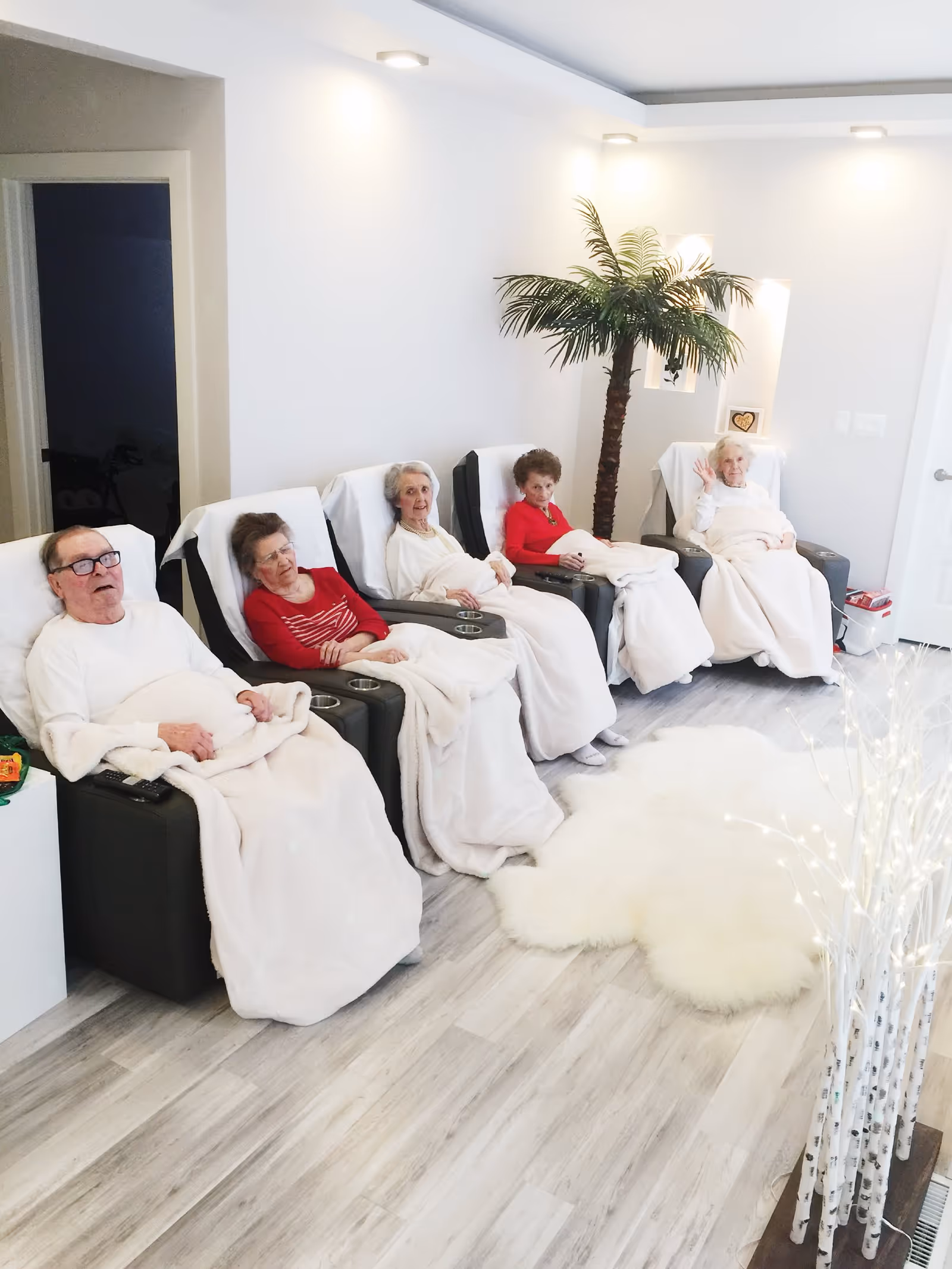 A group of five elderly people sitting in reclining chairs in a bright, modern room. Each person is covered with a light-colored blanket. The room has light wood flooring, white walls, a decorative palm tree, and a white fluffy rug on the floor. One elderly woman is waving at the camera.