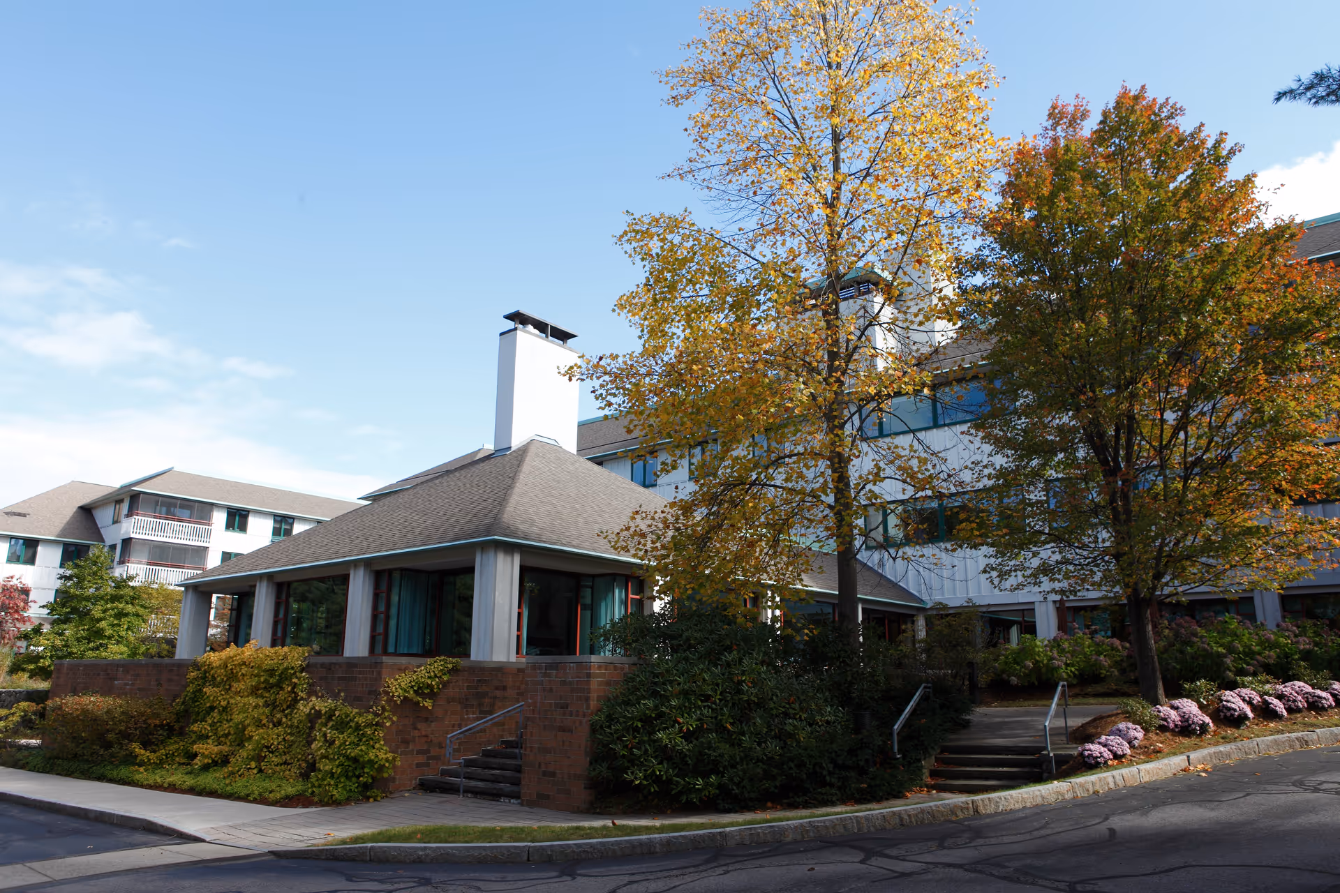 Exterior view of Orchard Cove facility showing a multi-story building with large windows, surrounded by trees with autumn foliage and landscaped bushes. There are stairs leading up to the entrance and a paved road in the foreground under a partly cloudy sky.