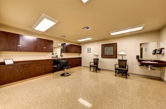 Interior view of a hair salon area in a senior living facility featuring two hair drying chairs, a salon chair in front of a large mirror, wooden cabinets along the wall, a wall-mounted sink with soap dispensers, and framed artwork on the walls.