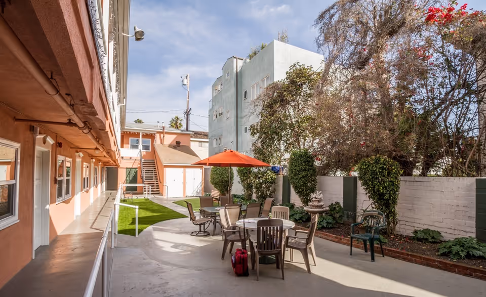 Sunlit outdoor courtyard with patio tables and chairs, an orange umbrella, walkways and surrounding buildings.