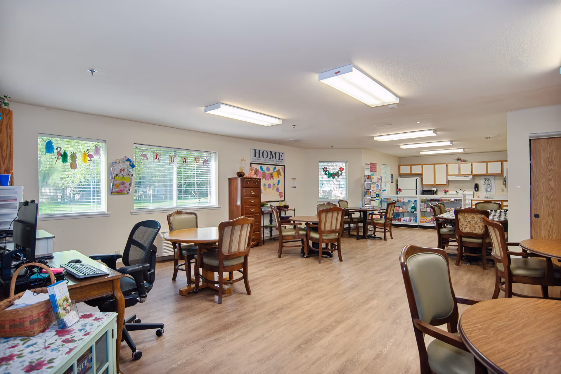 A spacious, well-lit common area in a senior living facility with multiple round tables and chairs arranged for socializing or activities. The room has large windows with blinds, a desk with a computer and office chair, and a kitchen area with cabinets and appliances in the background. Decorations and bulletin boards are visible on the walls.