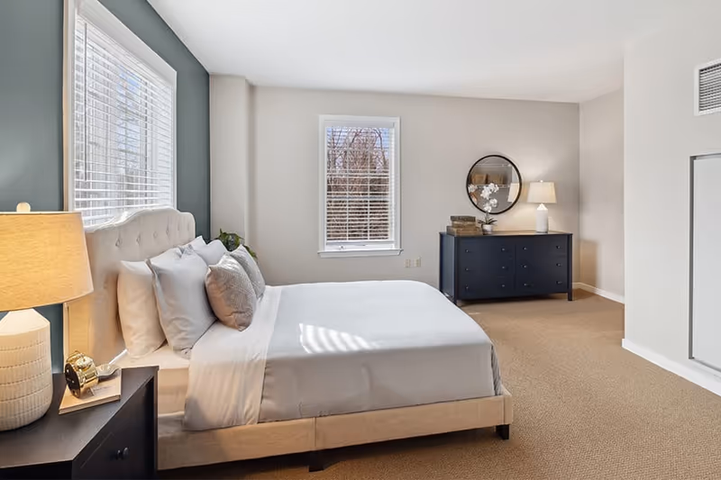 A bright and spacious bedroom with a large bed featuring white and gray pillows and bedding. There is a nightstand with a lamp and an alarm clock on the left side of the bed. The room has two windows with white blinds, a dark blue dresser with a round mirror above it, and a lamp on the dresser. The walls are painted in light neutral colors, and the floor is carpeted.