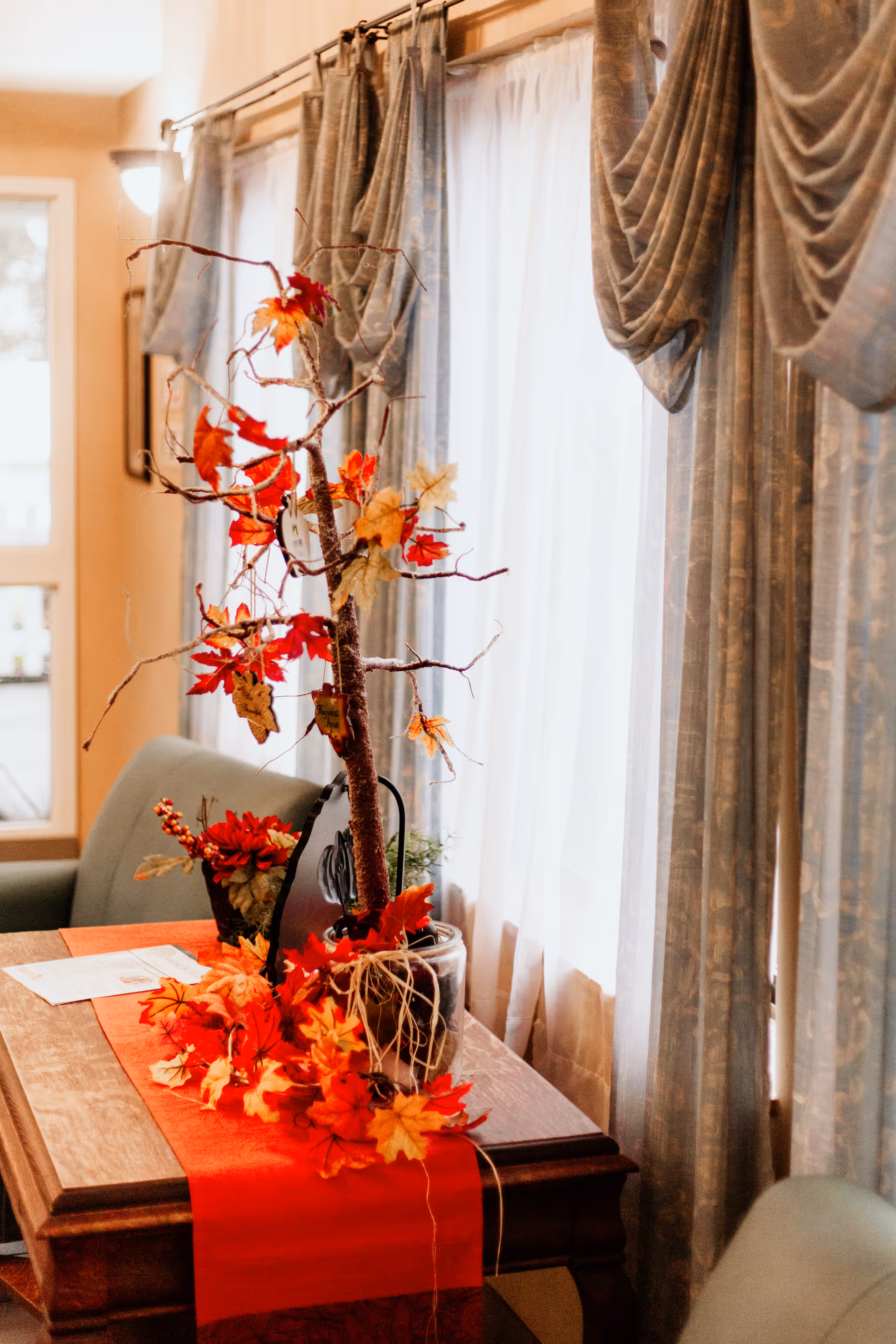 A cozy interior scene featuring a wooden table with a red table runner decorated with autumn leaves and a small artificial tree with red and yellow fall foliage. The table is positioned next to a window with sheer white curtains and heavy draped blue-gray curtains. Two green upholstered chairs are partially visible around the table.