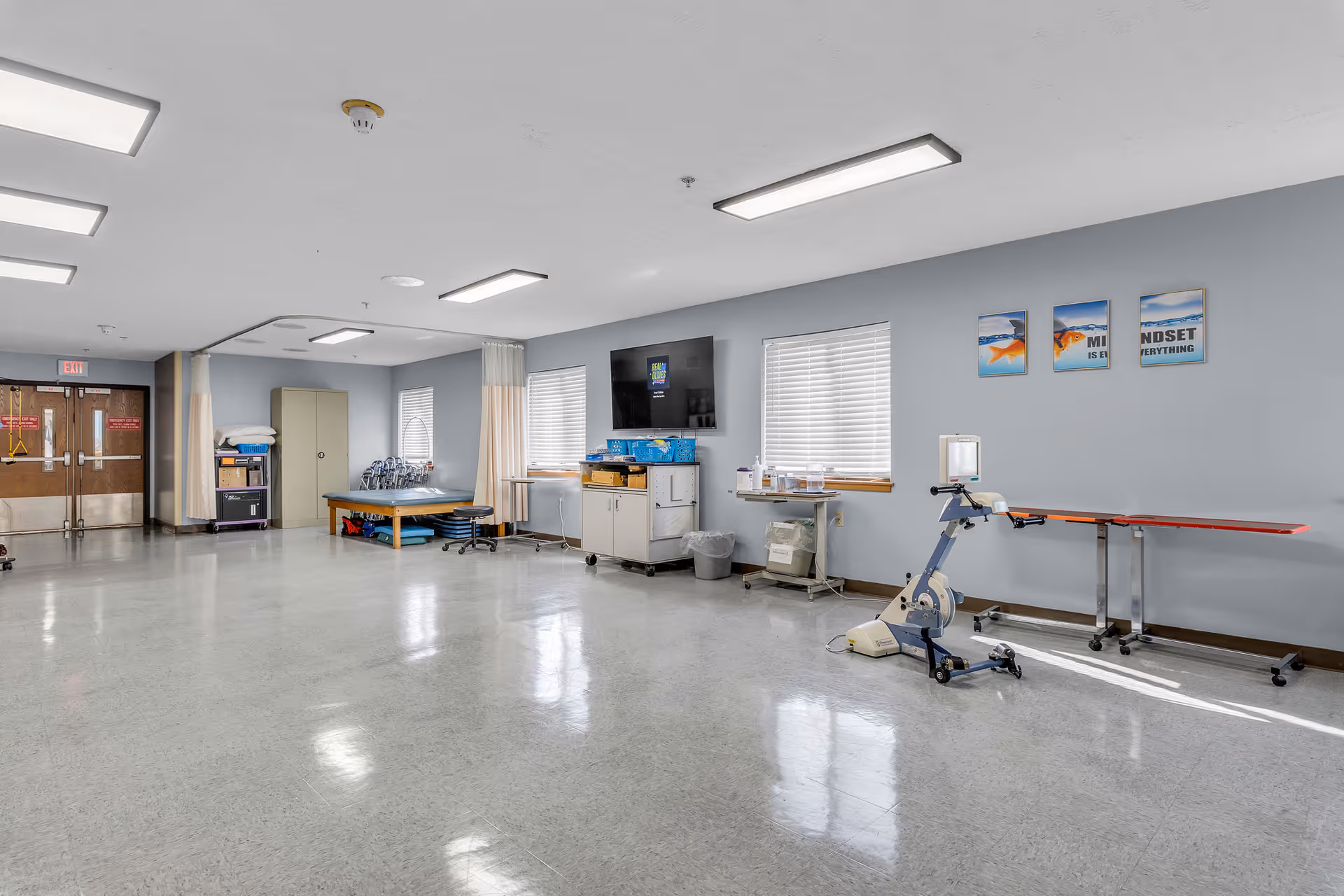 Spacious, well-lit rehabilitation/exercise room with therapy equipment, tables, a wall-mounted TV, and storage cabinets.