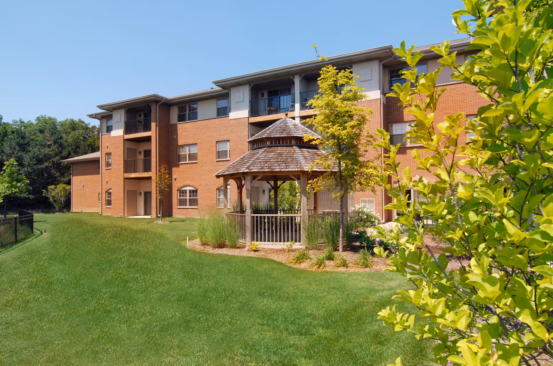 Outdoor view of a senior living facility with a three-story brick building in the background and a wooden gazebo surrounded by small trees and shrubs on a well-maintained grassy lawn under a clear blue sky.
