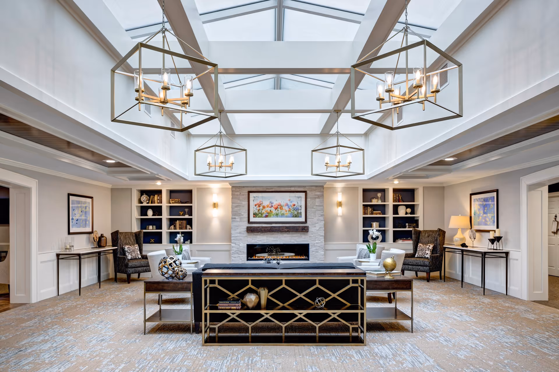 A spacious and elegant living room with a high ceiling and large skylights. The room features modern chandeliers, a stone fireplace with a colorful floral painting above it, built-in shelves with decorative items, and comfortable seating including armchairs and sofas. The room is decorated in neutral tones with accents of gold and dark wood furniture.