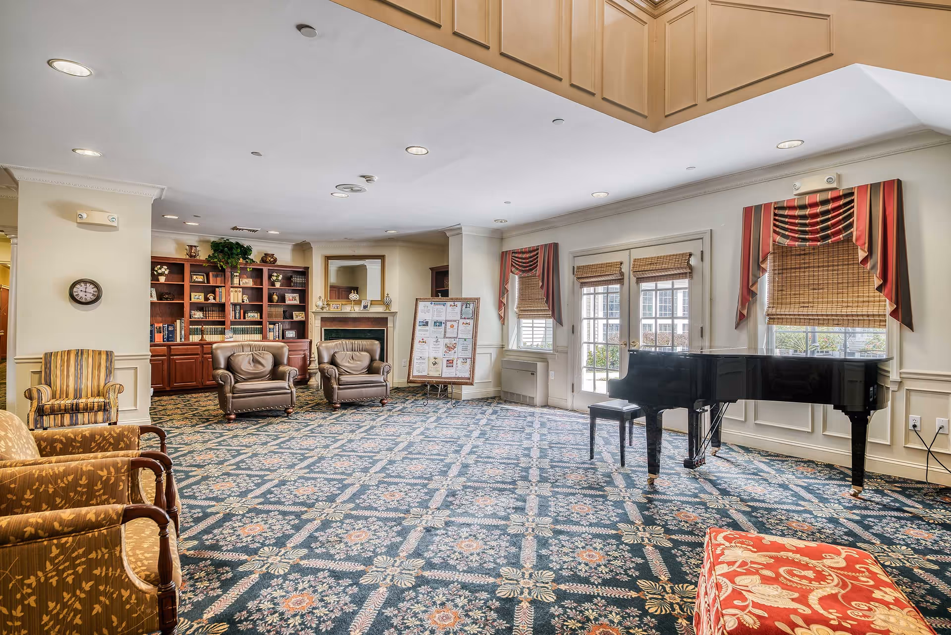 A spacious living room area with patterned carpet, two leather armchairs, a striped armchair, a red patterned ottoman, a black grand piano with a matching bench, and a wooden bookshelf filled with books and decorative items. There is a fireplace with a mirror above it, a bulletin board with notices, and windows with bamboo blinds and red striped valances letting in natural light.