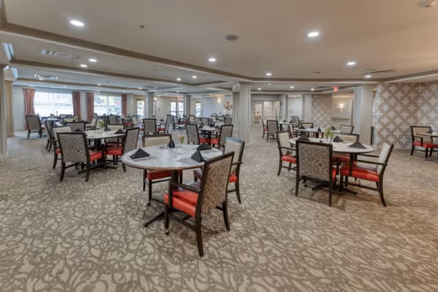 Spacious dining room with multiple round tables and chairs arranged neatly. Each table has place settings with black napkins and small floral centerpieces. The room has patterned carpet, large windows with curtains allowing natural light, and soft ceiling lighting.