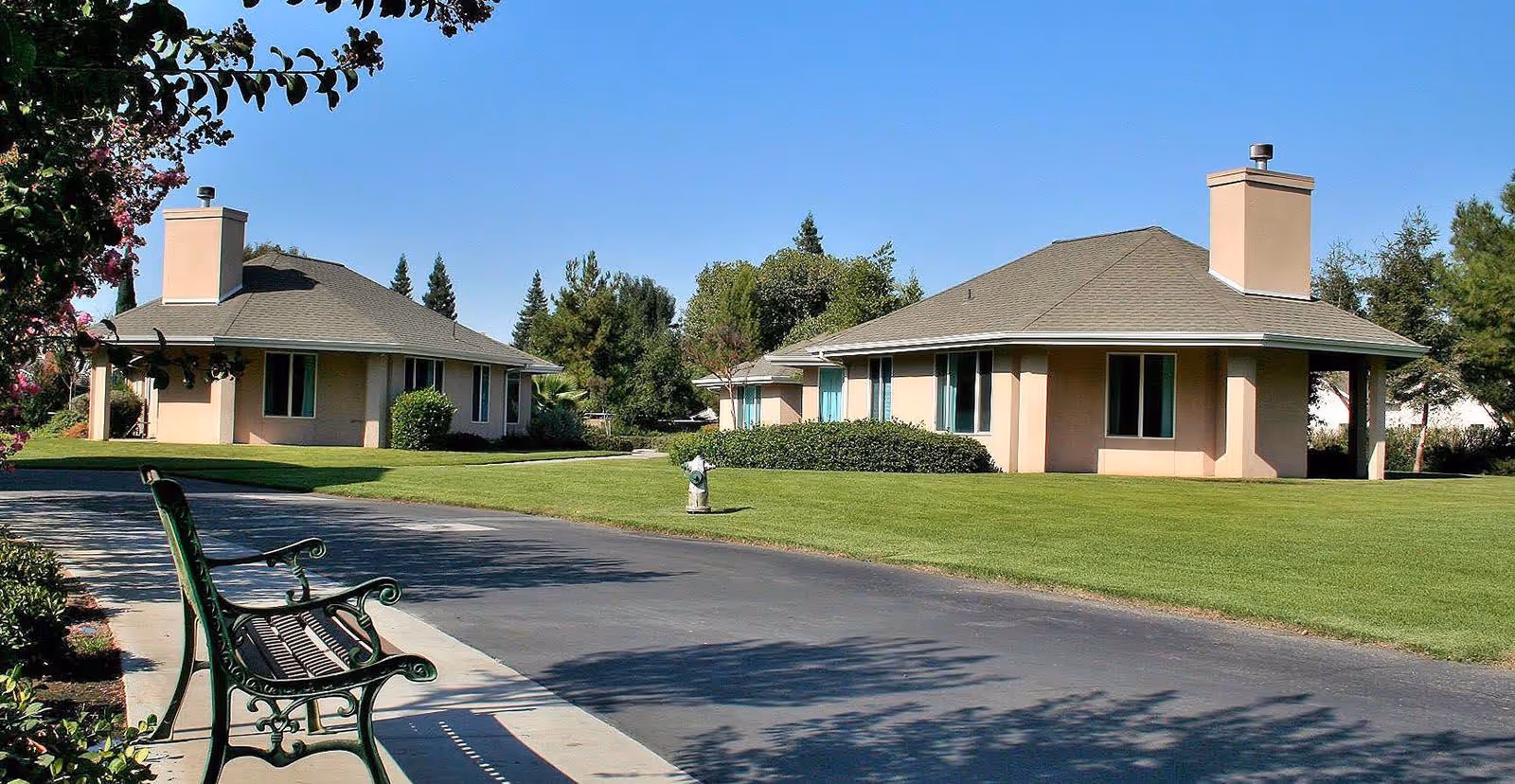 Single-story beige senior living cottages set on a manicured lawn with a bench and paved path in the foreground under a clear blue sky.