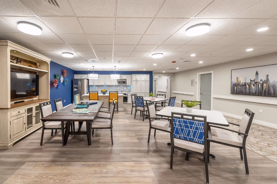 Bright communal dining and activity room with tables, chairs, a TV cabinet, and a kitchen area in the back.