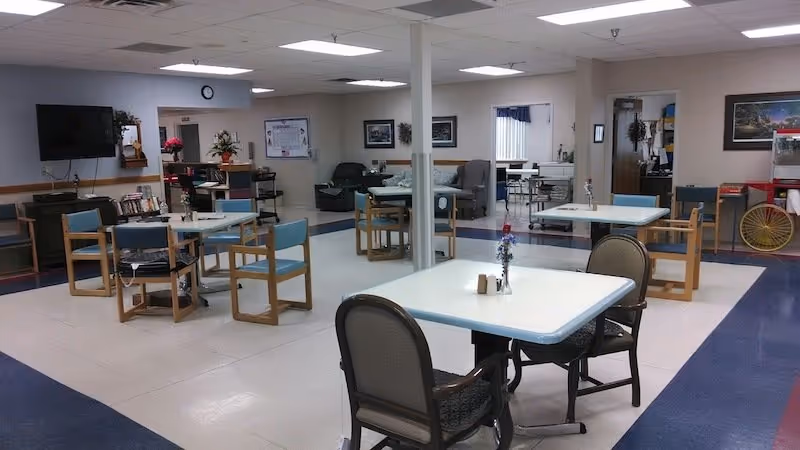 A spacious common area in a senior living facility with several tables and chairs arranged for seating. The room has a tiled floor with a blue border, a wall-mounted television, bookshelves, and some floral decorations. There are couches and additional seating in the background, with a popcorn machine visible on the right side.