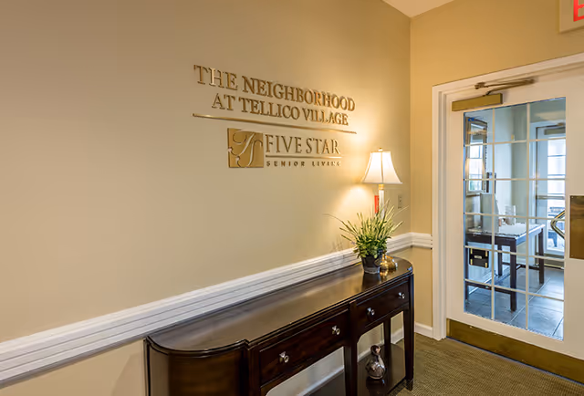 Interior hallway area with a dark wooden console table against a beige wall. Above the table, gold lettering on the wall reads 'THE NEIGHBORHOOD AT TELLICO VILLAGE' and 'FIVE STAR SENIOR LIVING' with a logo. A table lamp and a potted plant are placed on the console table. To the right, there is a glass door leading to another room with visible furniture inside.