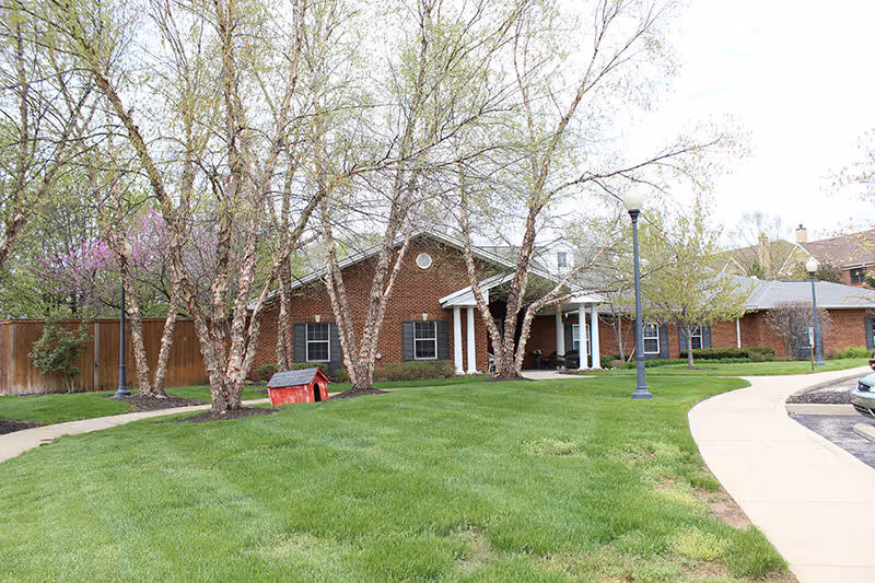 Single-story brick senior living building with a covered columned entrance, lawn, trees, and a curved walkway.