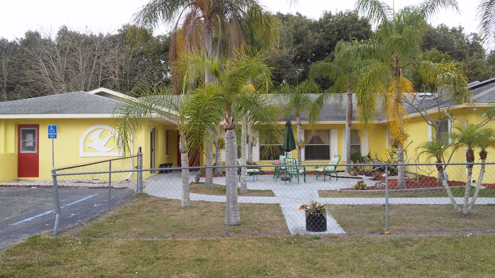Front exterior of a yellow assisted-living building with palm trees, a chain-link fence, a handicapped parking spot, and a small patio with green chairs and an umbrella.