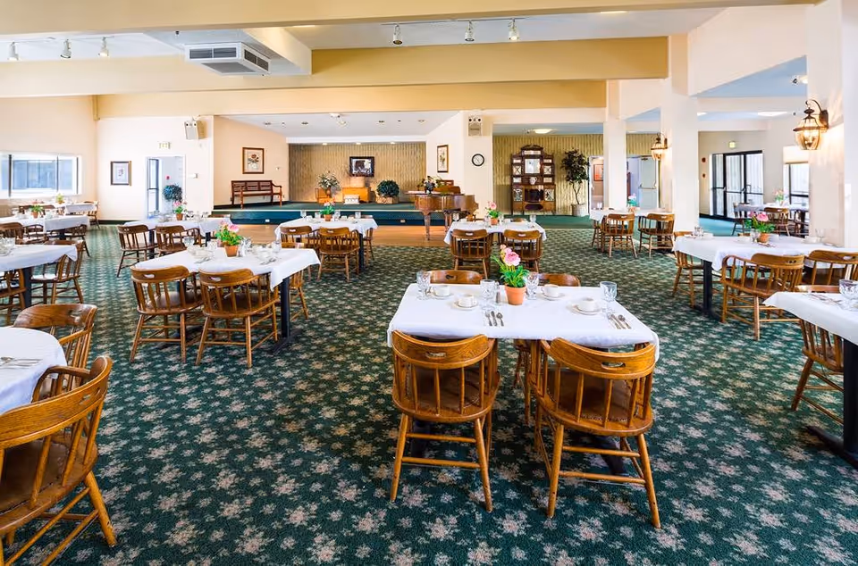 A spacious dining room with multiple wooden tables and chairs arranged neatly. Each table is covered with a white tablecloth and set with cups, saucers, glasses, and silverware. Small potted plants with flowers serve as centerpieces on the tables. The room has green carpet with a floral pattern, cream-colored walls, and large windows allowing natural light. In the background, there is a small stage area with a piano and some plants, along with framed pictures on the walls.