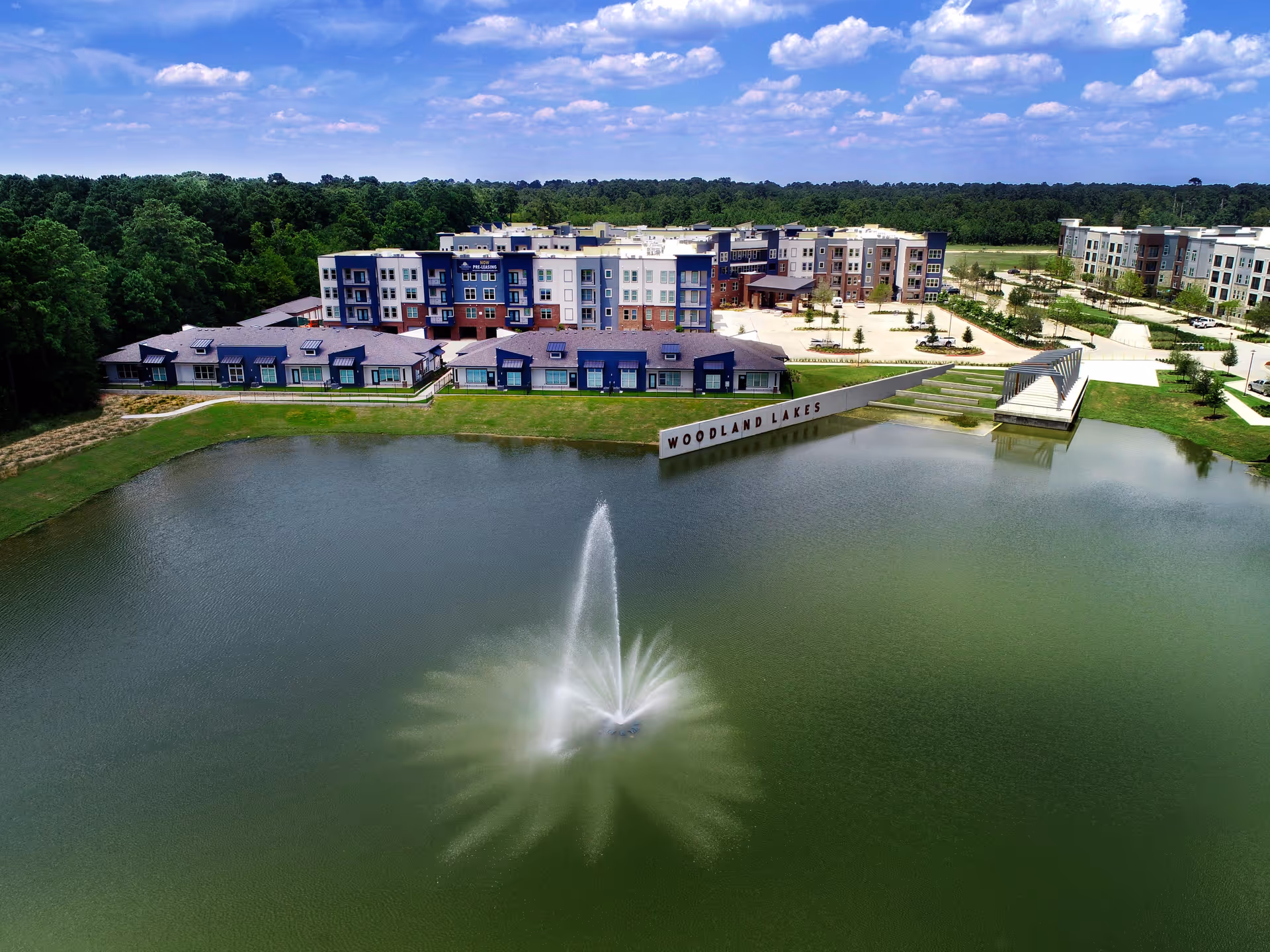 Aerial view of the Watermere at Woodland Lakes senior living facility showing multiple modern buildings surrounding a large pond with a central water fountain. The area is landscaped with trees and greenery under a partly cloudy sky.