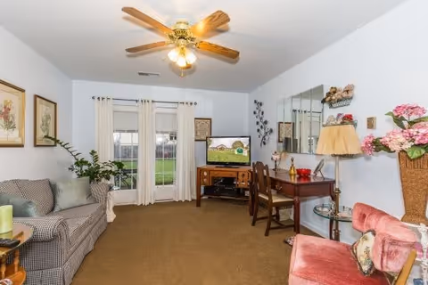 A cozy living room with a ceiling fan and light fixture. The room features a patterned sofa with cushions on the left, a wooden TV stand with a flat-screen television displaying a scenic image, and a wooden desk with two chairs on the right. There are decorative items including framed artwork on the walls, a large mirror, a table lamp, plants, and floral arrangements. The room has beige carpet flooring and white walls, with a window covered by sheer curtains letting in natural light.