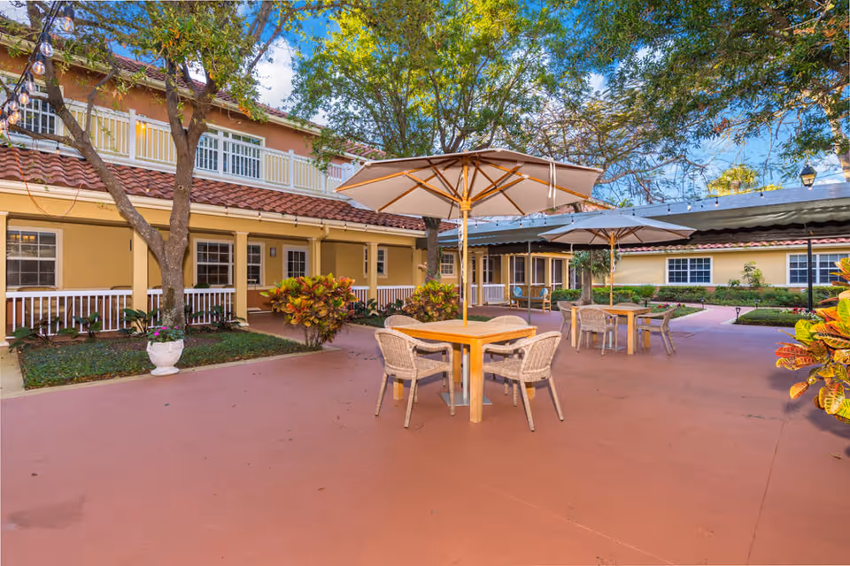 Outdoor courtyard area with several tables and chairs under large umbrellas, surrounded by a two-story building with yellow walls and white railings. Trees and plants add greenery to the space under a blue sky with some clouds.