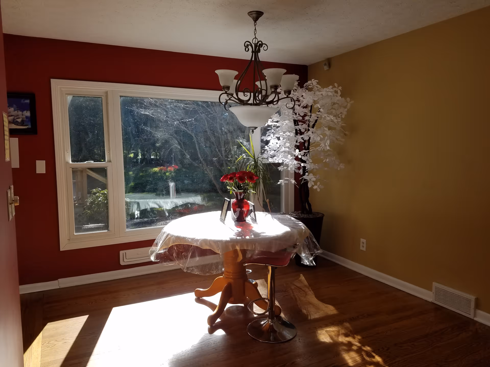 A small dining area with a round wooden table covered with a white tablecloth and a clear plastic cover. On the table is a red vase with red flowers and two small picture frames. The room has hardwood floors, a large window letting in sunlight, a red accent wall, and a yellow wall. A decorative chandelier hangs above the table, and there is a tall potted white artificial tree in the corner.