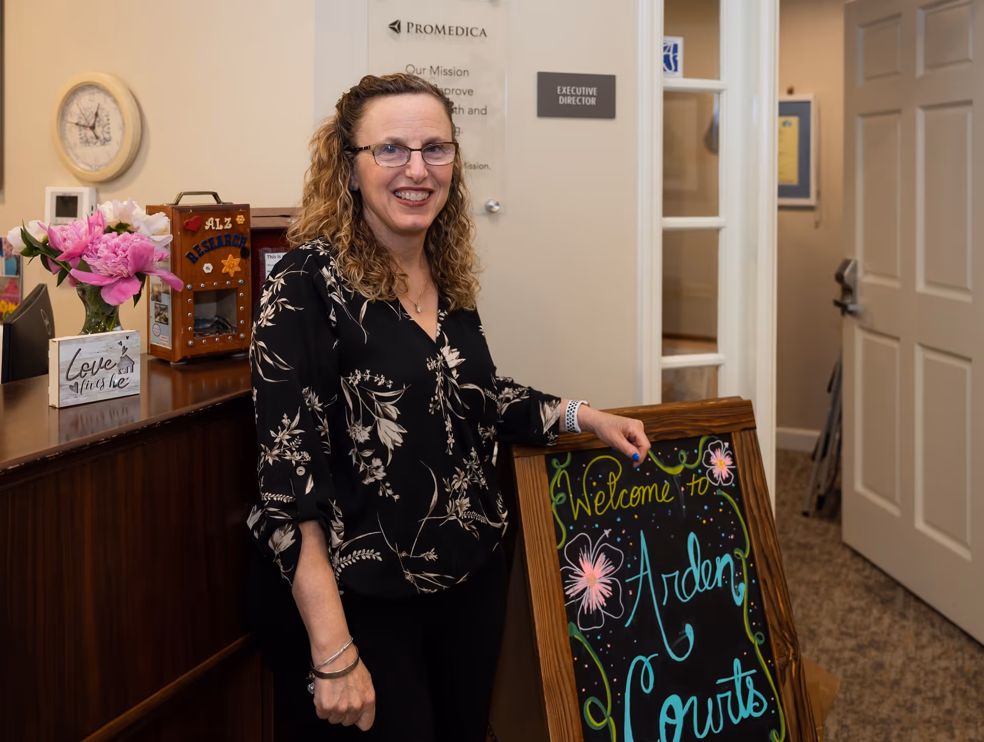 A woman with curly hair and glasses stands indoors next to a wooden counter and a decorative chalkboard sign that reads 'Welcome to Arden Courts.' Behind her is a door with a sign that says 'Executive Director' and a wall plaque with the ProMedica logo and mission statement. The counter has a vase with pink flowers and a small decorative sign that says 'Love lives here.'