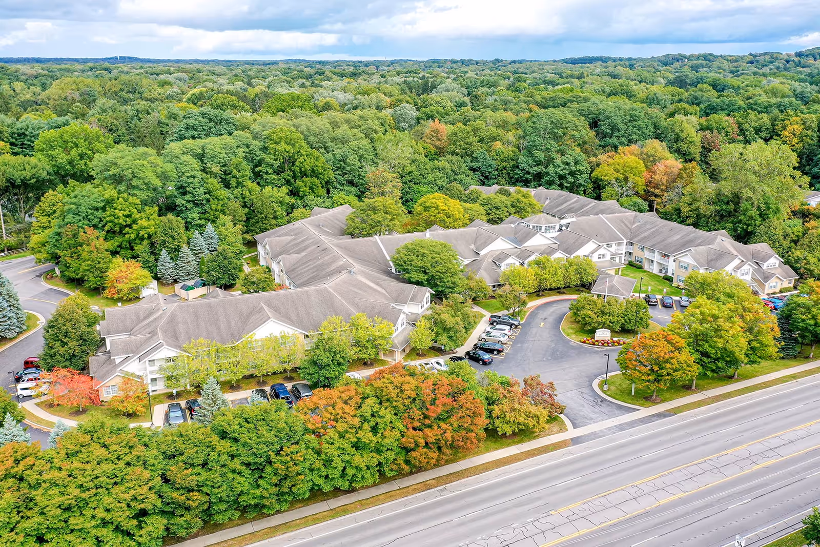 Aerial view of a large senior living complex surrounded by trees with parking areas and a road in front.