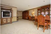 A spacious interior room with patterned carpet flooring, featuring a wooden dining table with chairs, a wooden cabinet with glass doors displaying decorative items, and a large wooden entertainment center with a television. The walls are painted beige with wooden trim, and the room is well-lit with ceiling lights.