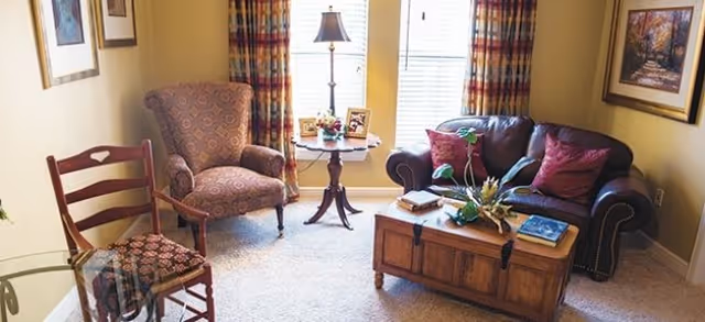 A cozy living room with a patterned armchair, a wooden chair with a cushioned seat, a dark leather sofa with red pillows, a wooden coffee table with decorative plants and books, a small round side table with a lamp and framed photos, and two windows with colorful curtains letting in natural light.