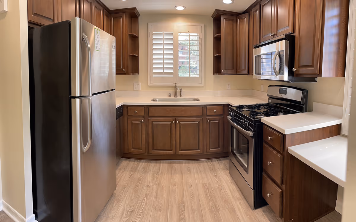 A modern kitchen with wooden cabinets, a stainless steel refrigerator on the left, a stainless steel gas stove with oven and a microwave above it on the right, and a sink centered under a window with white plantation shutters. The kitchen has light-colored countertops and wood flooring.