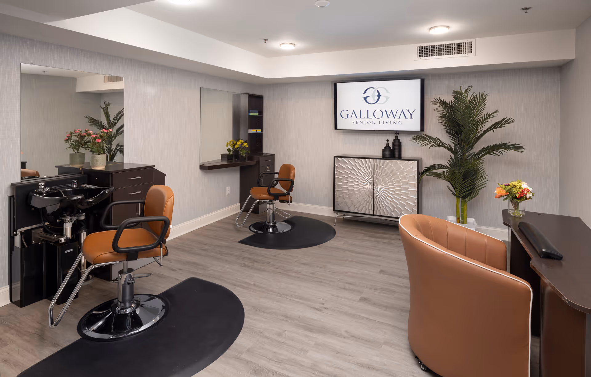Interior of a senior living facility hair salon with two brown salon chairs in front of mirrors, a black hair washing station, a decorative cabinet, plants, and a wall-mounted screen displaying the Galloway Senior Living logo.