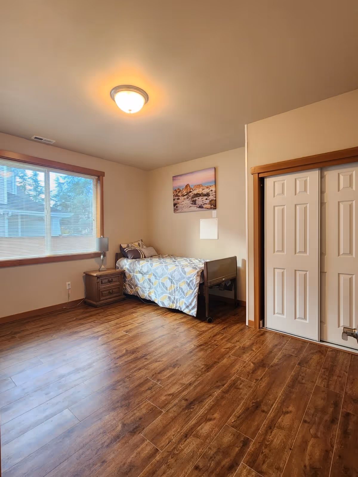 A simple bedroom with wooden flooring, a single bed with patterned bedding, a wooden nightstand with a lamp, a large window with blinds, a ceiling light fixture, a closet with white sliding doors, and a landscape picture hanging on the wall.