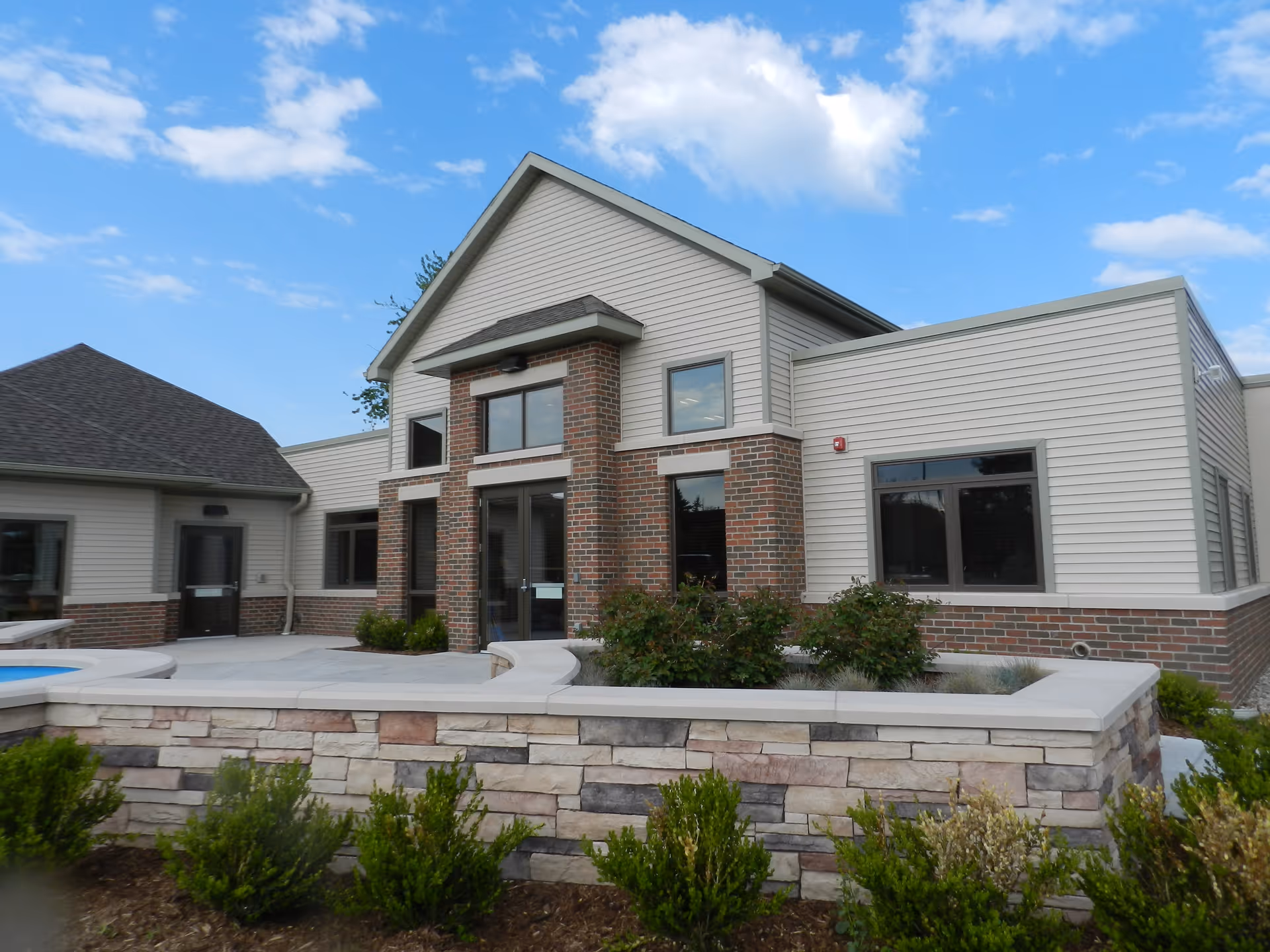 Exterior view of a single-story building with a combination of brick and light-colored siding. The building has multiple windows and a central entrance with glass doors. In front of the building is a low stone wall with some green shrubs and plants. The sky is partly cloudy with blue patches.