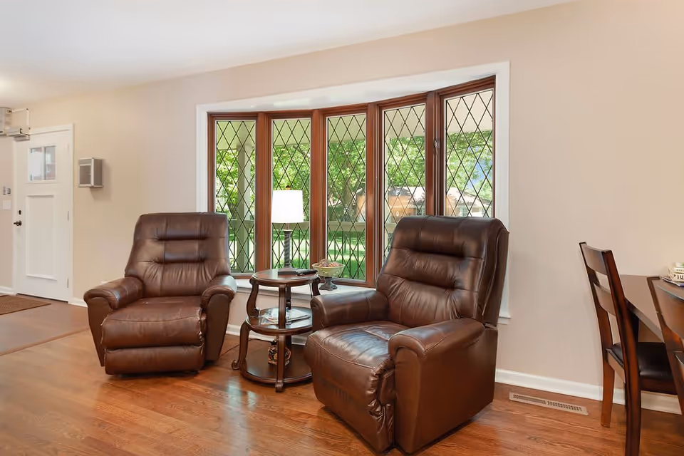 A cozy living room area with two brown leather recliner chairs positioned around a small round wooden side table with a lamp and decorative items. Behind the chairs is a large bay window with diamond-patterned glass panes, showing green trees outside. To the right, part of a wooden dining table and chairs is visible. The room has wooden flooring and light-colored walls.