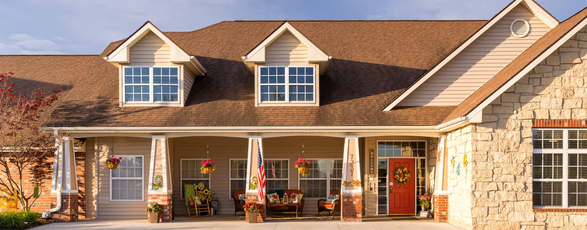 Front exterior of a senior living building with a covered porch, hanging flower baskets, an American flag, and a red door with a wreath.