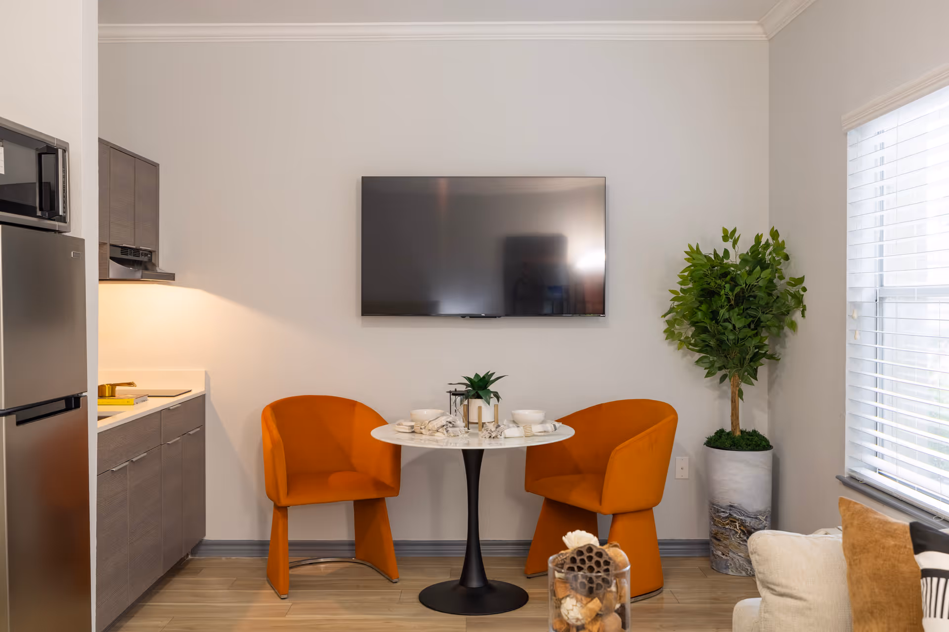 Cozy dining nook featuring two orange chairs at a round table beneath a wall-mounted TV, with a kitchenette to the left and a potted plant by a window.