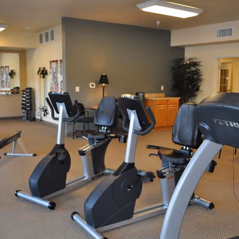 Interior view of a fitness room with exercise equipment including stationary bikes and a rowing machine. The room has beige carpet, a gray accent wall, a lamp on a small table, a plant in the corner, and a set of dumbbells on a rack near a mirror.