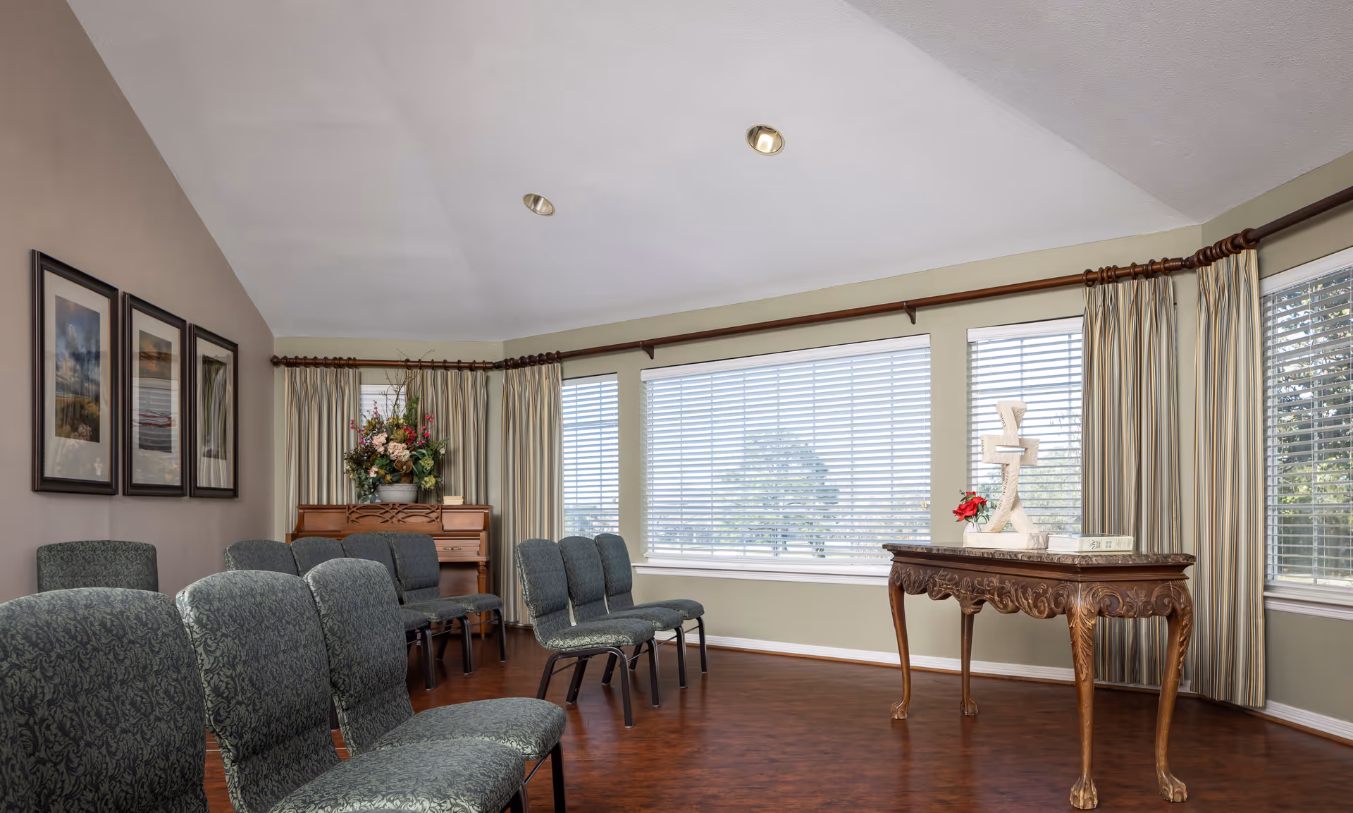 A quiet room with rows of green upholstered chairs arranged facing a wooden table with decorative items, including a cross and flowers. The room has large windows with blinds and curtains, a piano with a flower arrangement on top, and framed pictures on the wall.