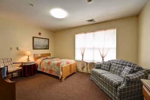 A cozy bedroom with a single bed covered in a patchwork quilt, a wooden headboard, a side table with a lamp, a framed painting on the wall, a plaid upholstered sofa, a small round table near a window with sheer curtains, and a chair with a patterned cushion.