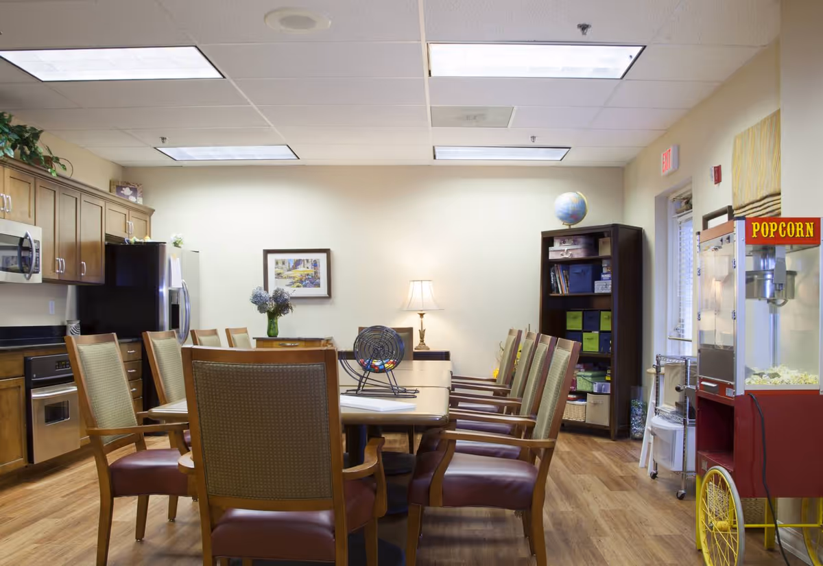 A communal dining and activity room with a long table surrounded by wooden chairs with cushioned seats. The room features a kitchen area with wooden cabinets, a microwave, an oven, and a refrigerator. On the right side, there is a popcorn machine filled with popcorn. A bookshelf with various items and a globe is against the far wall, along with a table lamp and a framed picture on the wall. The room has a wooden floor and ceiling lights.