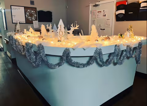 Reception desk decorated with silver tinsel garlands, white Christmas lights, and white holiday-themed ornaments including trees, reindeer, and a sign spelling JOY in a healthcare facility.