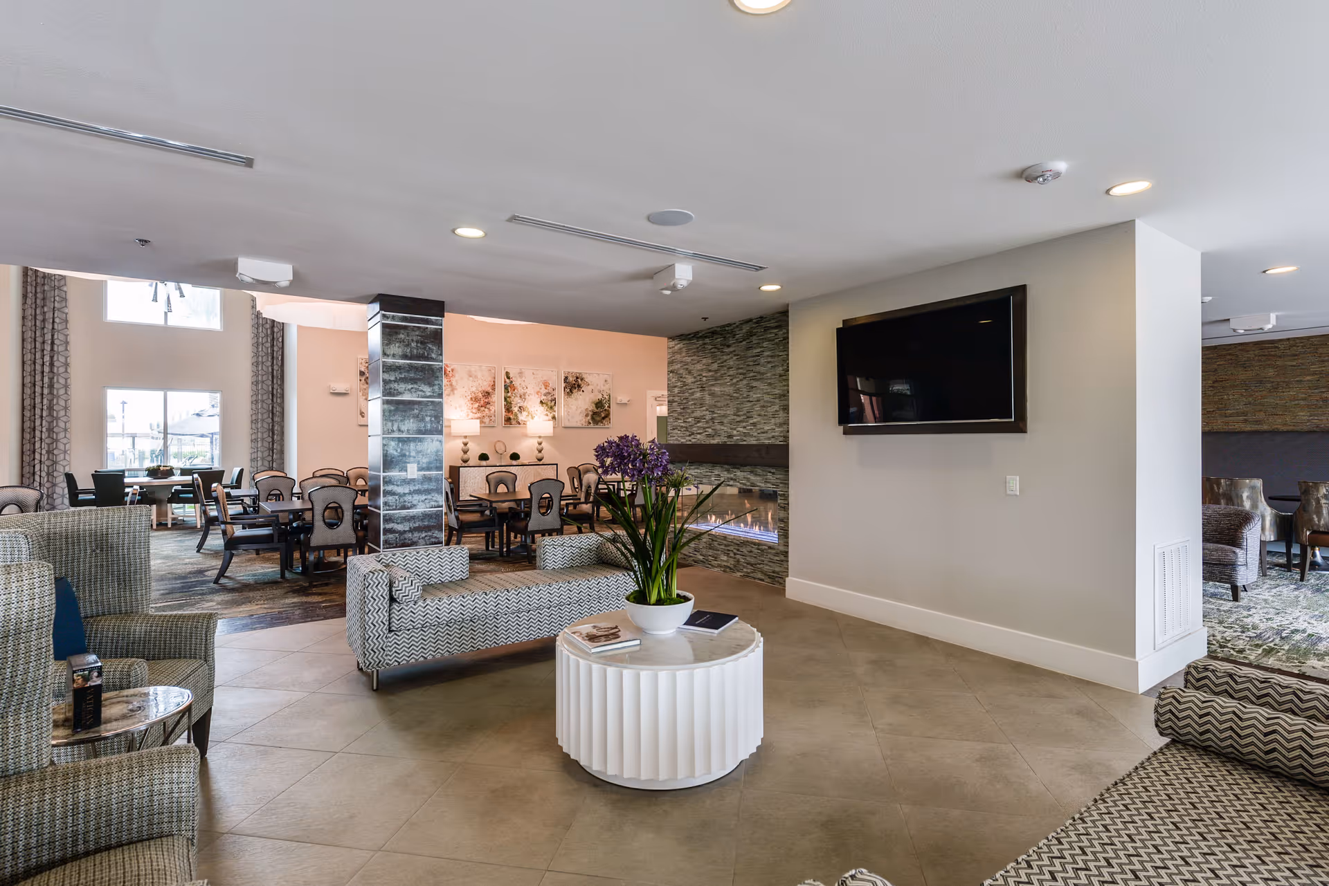 A modern senior living facility common area with patterned armchairs and sofas arranged around a white round coffee table with a potted plant. In the background, there is a dining area with tables and chairs, a wall-mounted TV, and a contemporary fireplace with a stone surround. Large windows allow natural light to fill the space.