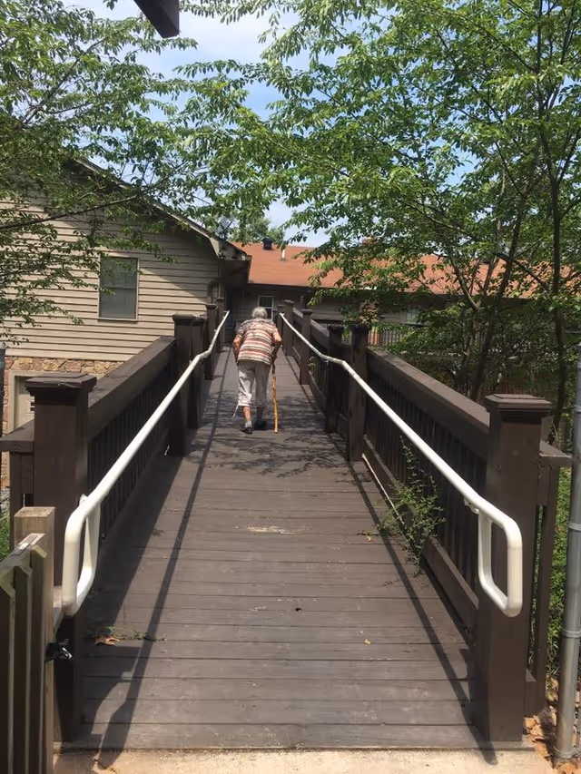 An elderly person walking up a wooden ramp with white handrails on both sides, surrounded by trees and leading to a building with beige siding and a red roof.