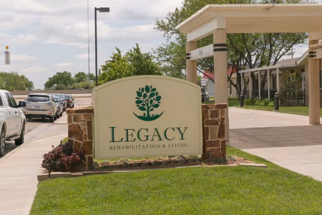 Outdoor view of the entrance area of Legacy Rehabilitation and Living facility, featuring a large sign with the facility's name and logo, a covered drop-off area, parked cars along the street, and green grass with some landscaping.