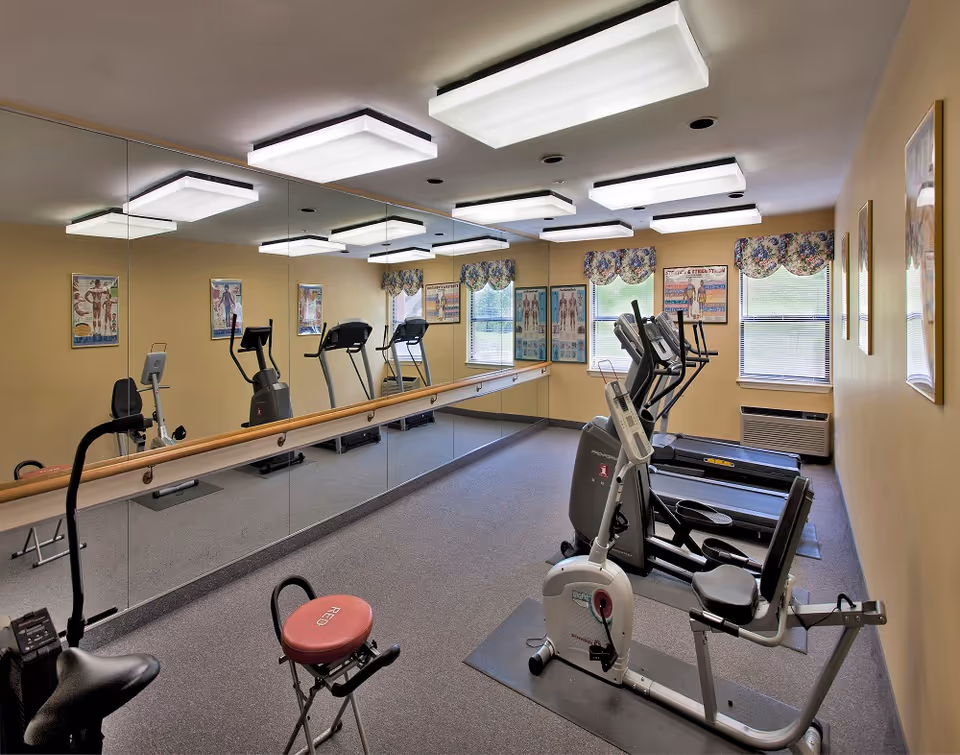 Small fitness room with treadmills, stationary bikes, mirrors and a ballet barre under rectangular ceiling lights.