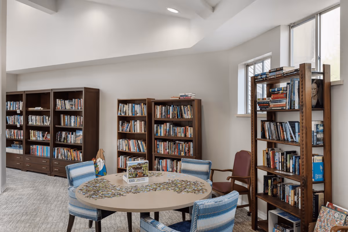 A cozy reading and puzzle area in a senior living facility featuring multiple wooden bookshelves filled with books, a round table with a partially completed 1000-piece puzzle, and several upholstered chairs. The room has light-colored walls and carpeted flooring, with windows letting in natural light.