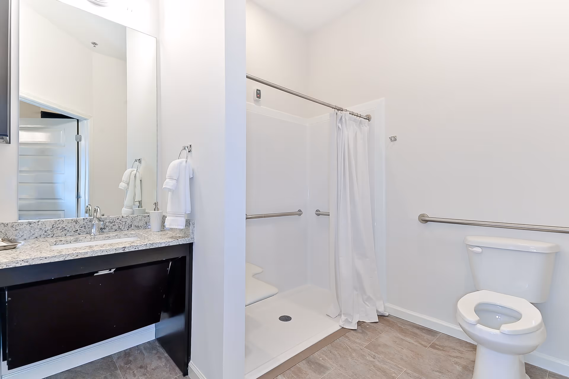 Bright accessible bathroom with a granite-topped vanity and mirror, walk-in shower with curtain and grab bars, and a toilet with a safety rail.