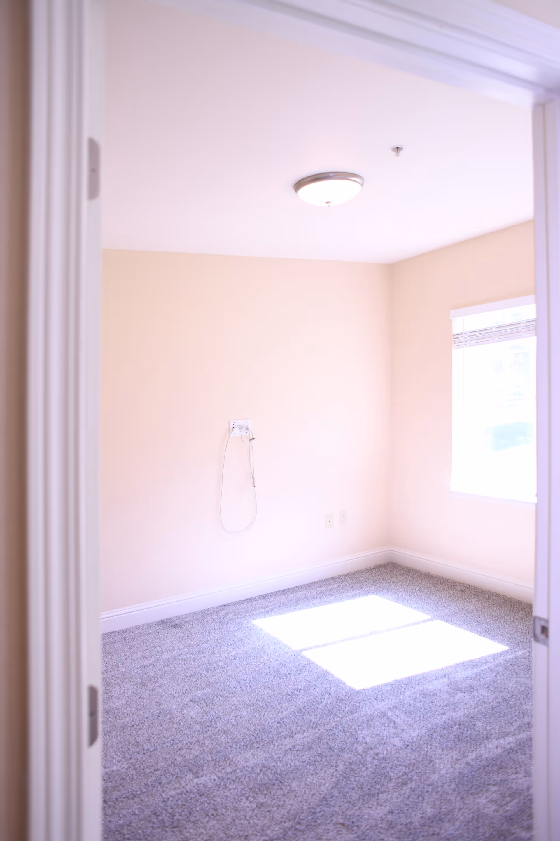 Empty room with beige walls, gray carpet, a ceiling light fixture, and a window letting in sunlight. There is a wall outlet with a cable hanging on the wall.