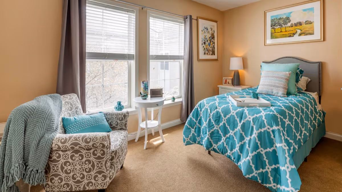 A cozy bedroom with a single bed covered in a turquoise and white patterned comforter and multiple pillows. Next to the bed is a white nightstand with a lamp and a framed picture. There is a patterned armchair with a turquoise pillow and a matching throw blanket draped over it. Two large windows with gray curtains let in natural light. A small round white table with decorative items is placed between the windows. The walls are painted beige and decorated with framed artwork.